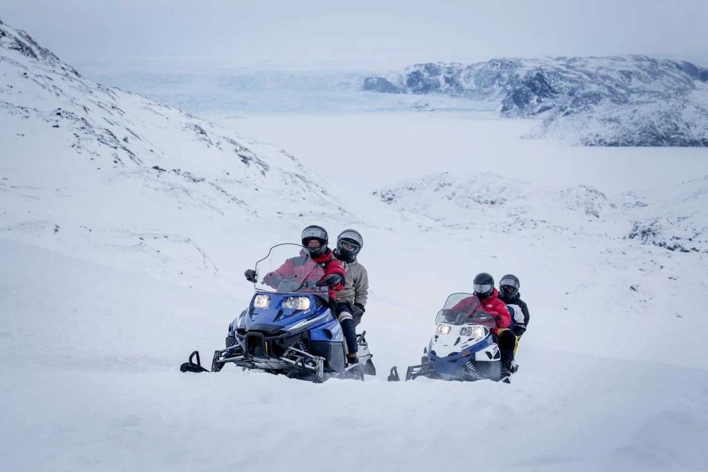 A group of people, wearing helmets and winter gear, ride snowmobiles through a vast, snow-covered landscape. A large frozen bay and mountains are visible in the background.