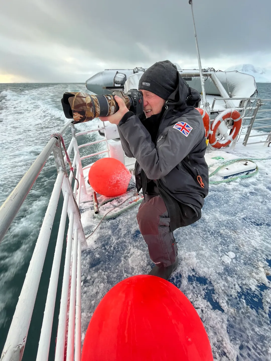 Tour guide Conrad Dickinson kneels on a snow-covered boat deck, aiming a large camera with a camouflage lens cover. He wears a dark jacket with a Union Jack patch on the sleeve.