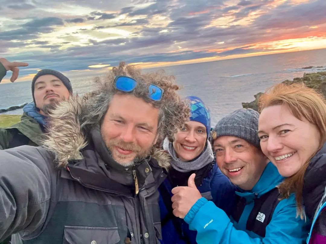 A group of five people smiling and posing for a selfie with the sea and a sunset sky in the background.