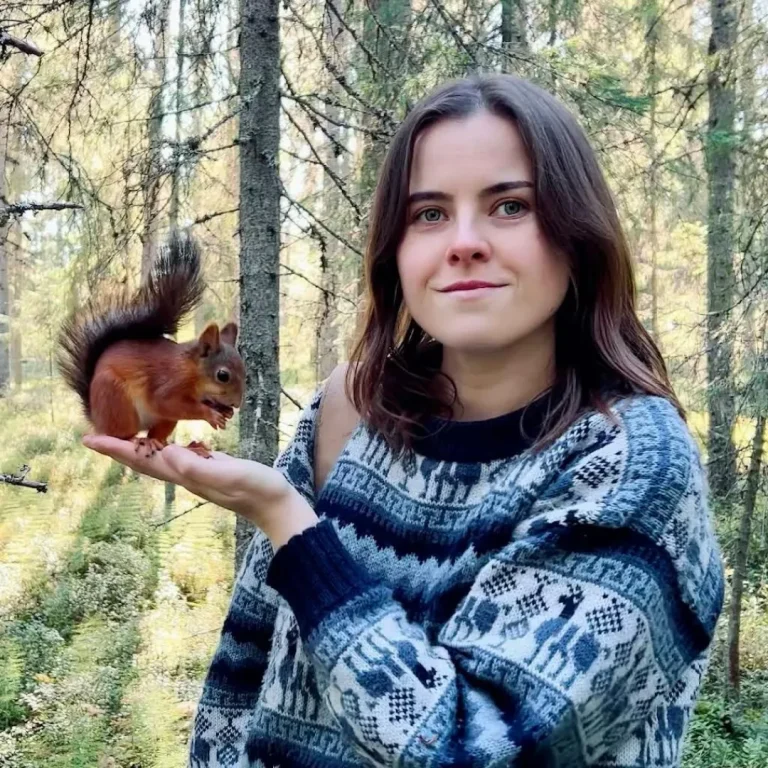 Tour guide Dani Connor stands in a forest holding a small red squirrel in her open hand while smiling at the camera.