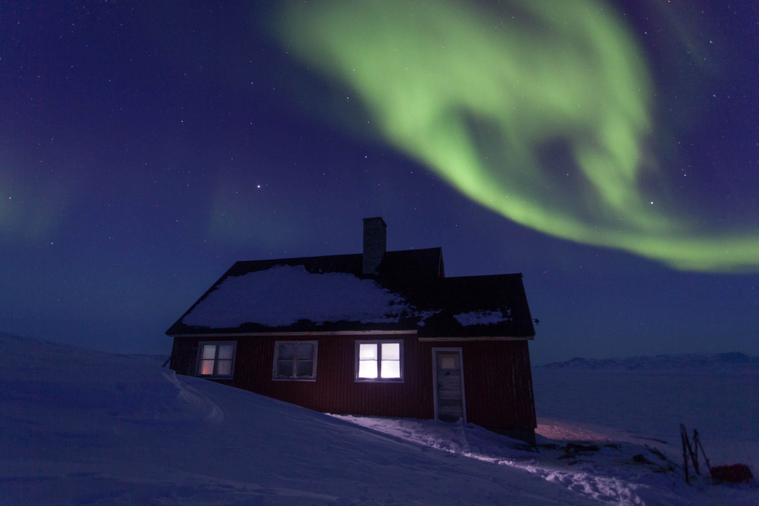 A small, rustic red cabin with a snow-covered roof sits on a snowy hill at night - a bright green band of Northern Lights arcs across the starry sky above it.