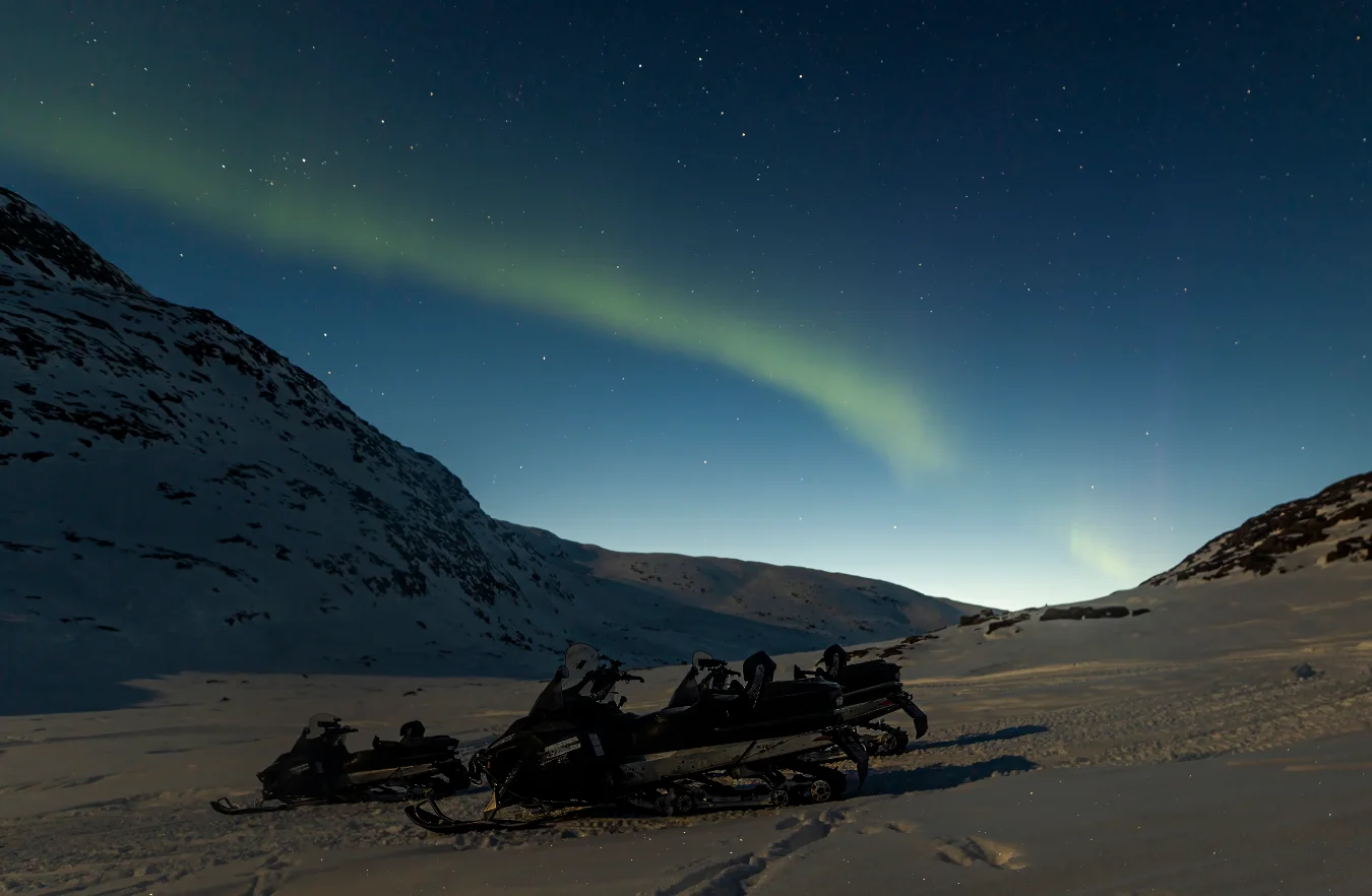 Three snowmobiles are parked in a snowy valley at night. Above them, a bright green band of Northern Lights arcs across a dark blue sky filled with stars.
