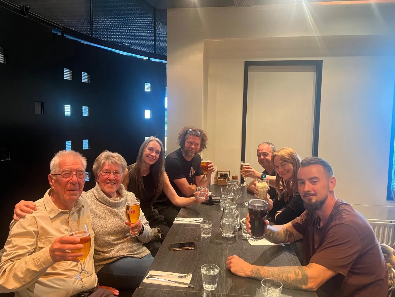A group of seven smiling clients and guides sitting around a long dining table, raising their glasses and drinks for a toast.