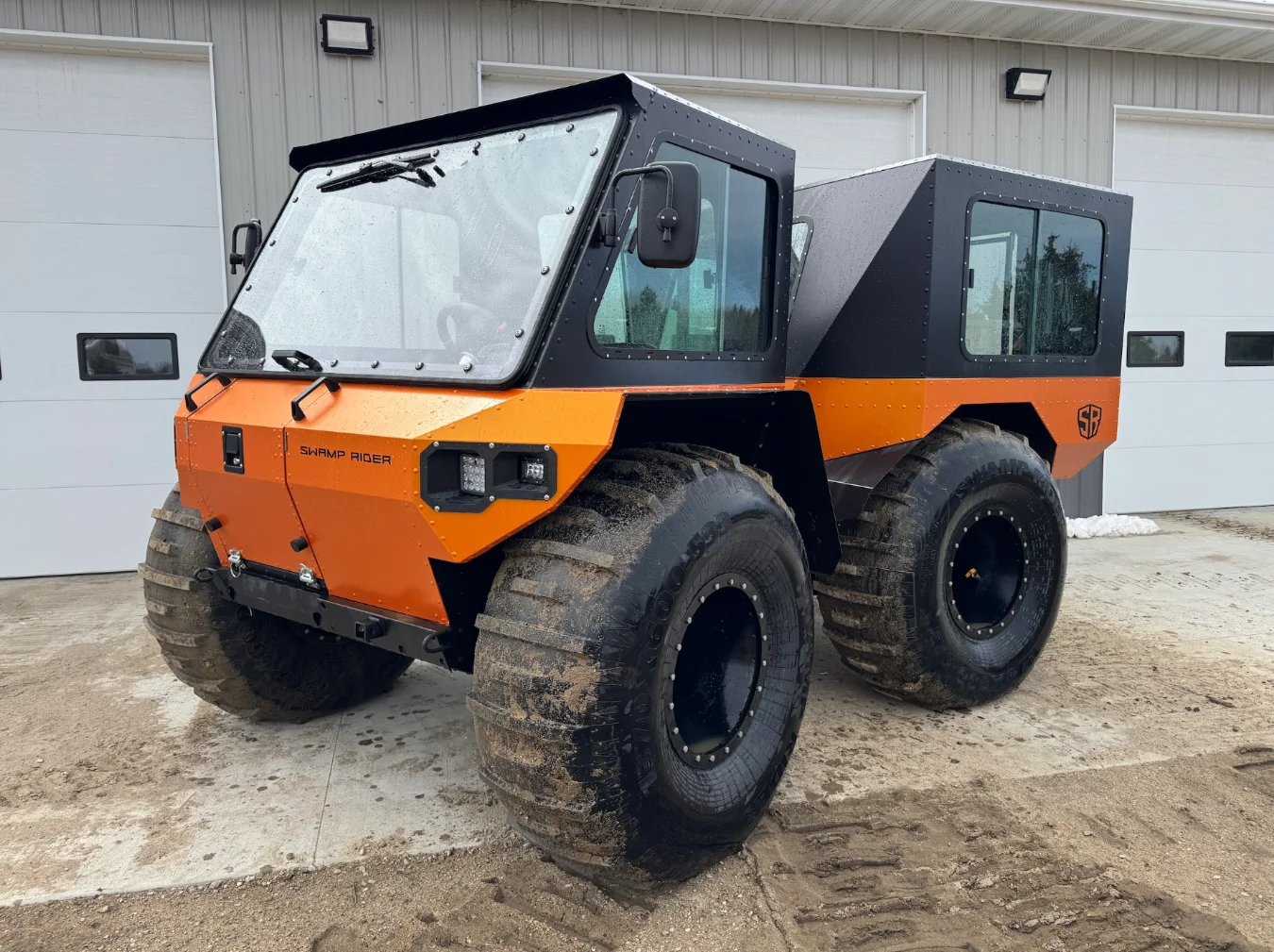 A large orange-and-black all-terrain vehicle with huge muddy tyres, parked on a dirt and gravel surface in front of a white building with garage doors.
