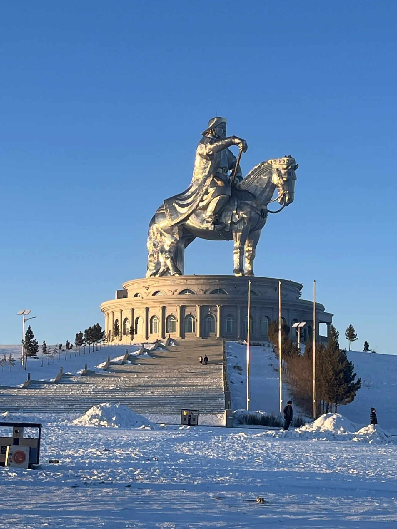 The Genghis Khan Equestrian Statue, a massive gleaming silver monument of a rider on a horse, situated atop a columned circular building in a snowy landscape under a clear blue sky.