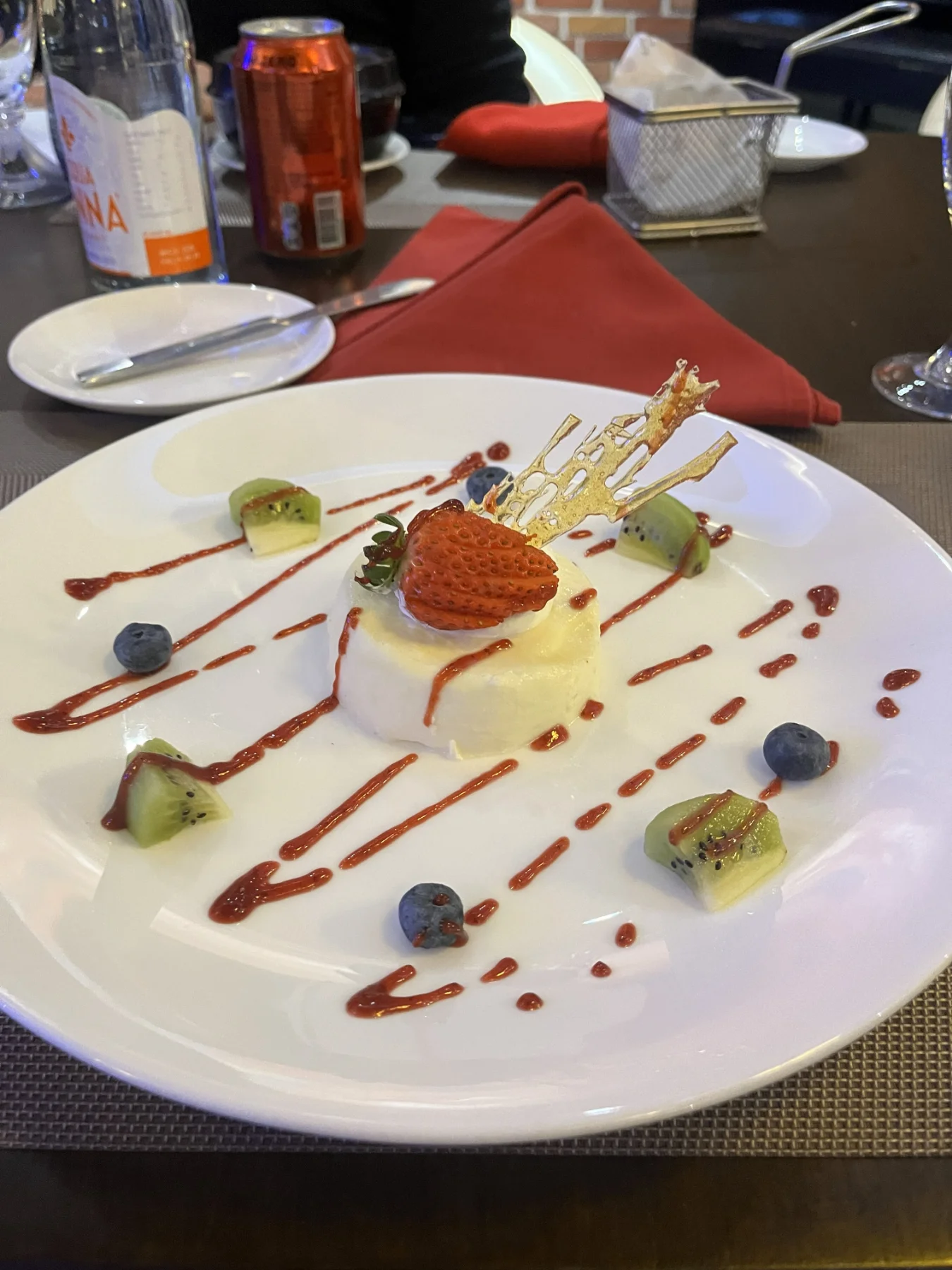 An elegantly plated dessert featuring a small round white cake topped with a fresh strawberry and a delicate, lattice-like sugar garnish. The white plate is decorated with diagonal drizzles of red sauce, whole blueberries and slices of kiwi. A red napkin and various drinks are also visible on the table.