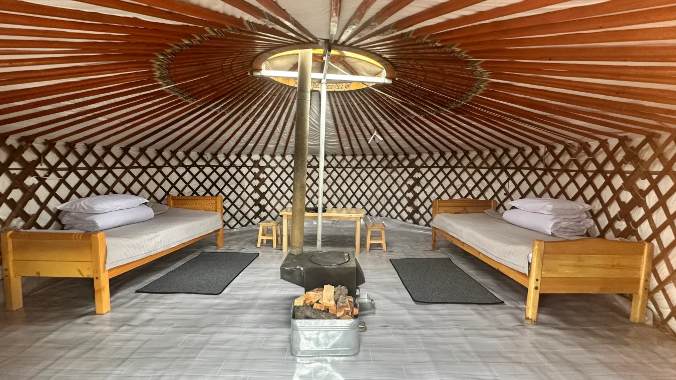 The interior of a traditional Mongolian ger featuring two wooden single beds with white bedding, a central metal stove with firewood, a small wooden table, and latticed wooden walls.